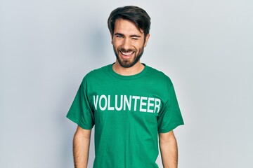 Young hispanic man wearing volunteer t shirt winking looking at the camera with sexy expression,...