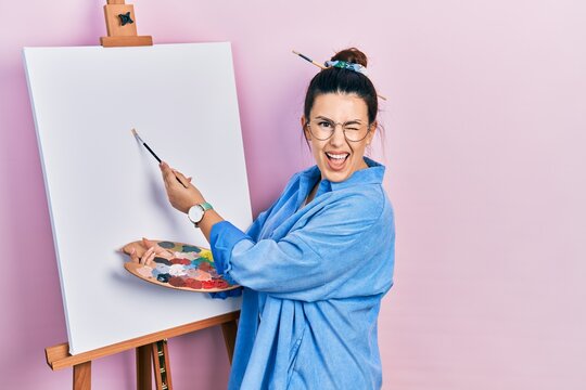 Young hispanic woman standing drawing with palette by painter easel stand winking looking at the camera with sexy expression, cheerful and happy face.