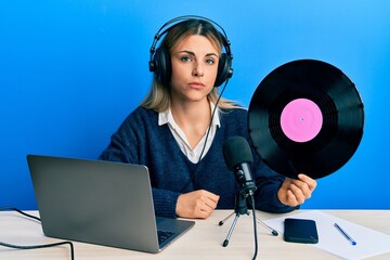 Young caucasian woman working at radio studio holding vinyl disc thinking attitude and sober expression looking self confident