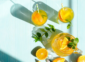 Glasses of lemonade with lemons, oranges and mint leaves on blue wooden background. Top view of lemonade. Summer cold drinks under direct sun shines. Caustic reflection effect.
