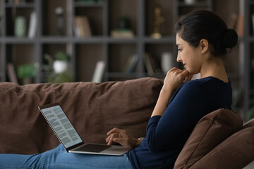 Happy millennial Indian woman sit relax on couch use computer study online from home. Smiling young...
