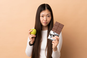 Young Chinese girl over isolated background taking a chocolate tablet in one hand and an apple in...