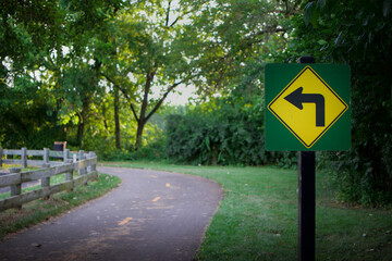 Yellow left turn ahead warning sign on bike path with green trees nature