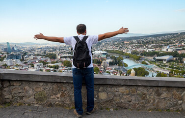 Rear view of tourist with backpack rise his hands and looking to the old city of Tbilisi and enjoying view - obrazy, fototapety, plakaty