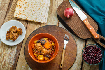 Jewish dish chelnt with meat on the table in a plate near ingredients and matzo.