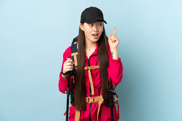 Young Chinese girl with backpack and trekking poles over isolated blue background intending to realizes the solution while lifting a finger up