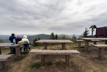 Wielki Lubon, Poland: Bunch of people sitting on a wooden bench at a peak of polish mountain during cloudy rainy weather located in a region of Beskid Wyspowy