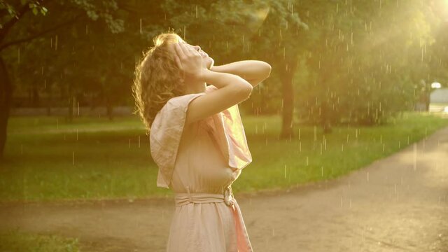 a woman in a dress, enjoys the rain in the light of the departing sun, she raises her hands and covers her face with happiness, the image of a beautiful youth, youth of the soul