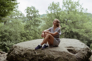Young woman in summer dress sitting on a big rock in the forest, having rest or meditating