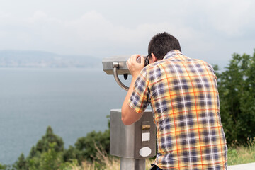 Obraz premium young man using stationary binoculars on a warm summer day on a hill against the background of the mountains and sea