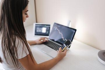 Attractive young woman sits at table in front of laptop, tablet and uses smartphone for work. Beautiful brunette girl analyzes stock market while at home