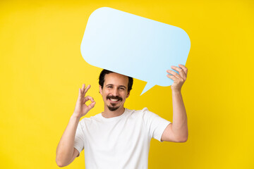 Young caucasian man isolated on yellow background holding an empty speech bubble and doing OK sign