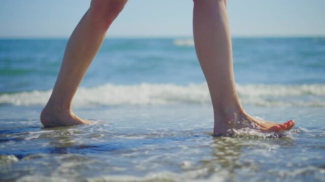 Close-up side tracking footage of woman legs being washed by ocean waves while walking on beach. Summer vacation at sea, relaxing on beach.