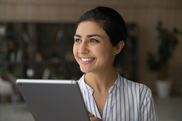 Smiling millennial Indian woman use tablet gadget look in distance thinking planning. Happy young mixed race female hold pad device talk speak on webcam video call. Communication, technology concept.