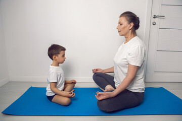 grandmother and her grandson are sitting on a yoga mat in a white apartment at home and meditating in the lotus position