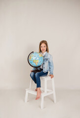 a little blonde girl in denim is sitting with a globe on a white background with a place for text. Back to school