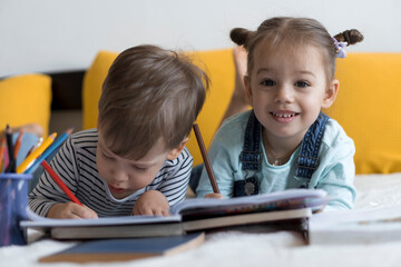 smart smiling preshool toddler children boy and girl draw with pencils lying on their stomach at yellow bed. little cute reader have fun, happy kid on quarantine at home. Friendship, family, education