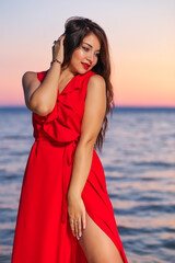 Beautiful brunette girl in a red dress posing on the seashore during sunrise or sunset