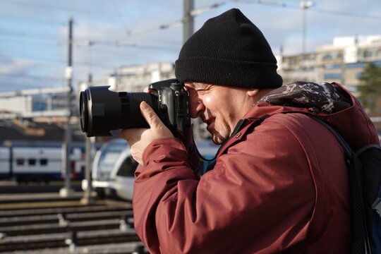 A man taking photos of trains in winter. Side view photo. He is wearing a cap and winter jacket. 
