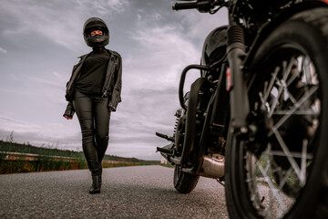Sexy woman in helmet sits goes to her classic black motorcycle. Female confident motorcyclist alone on highway. Ready for a trip. Cafe racers, motorbike aesthetics and vintage design concept.