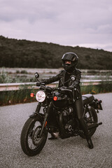 Fototapeta na wymiar Stylish motorcyclist woman in helmet and leather jacket sitting on vintage motorcycle. Female driver outdoors on nature background. Trip, cafe racers, speed, freedom concept.
