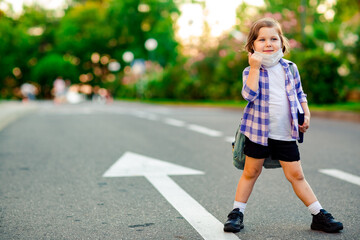 a schoolgirl is standing on the road in a plaid shirt and a medical mask, with a school backpack and a diary in her hands