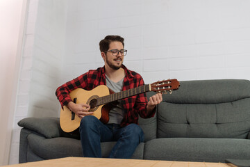Latin hipster in glasses and red plaid shirt tuning the guitar on the couch of a house.
