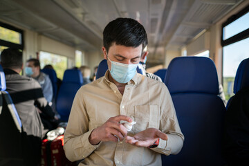 Travel safely on public transport. Young man with face mask using wash hand sanitizer gel dispenser. Passenger with protective mask disinfects hands in train carriage. Masked commuter use alcohol gel