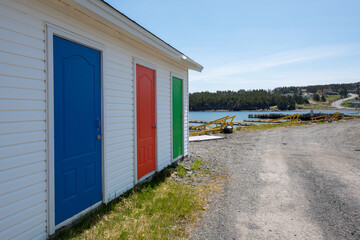 Fototapeta na wymiar A row of colorfully painted solid doors of blue, red, and green. The exterior wall is white vinyl siding. The sky is blue in the background and the storage units are sitting on gravel with blue ocean.
