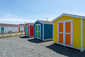 Obraz na płótnie Canvas A row of small colorful painted huts or sheds made of wood. The exterior walls are colorful with double wooden doors. The sky is blue in the background and the storage units are sitting on gravel.