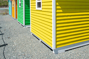 A row of small colorful painted huts or sheds made of wood. The exterior walls are colorful with double wooden doors. The sky is blue in the background and the storage units are sitting on gravel.