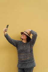 Vertical image of a woman taking her hat and taking a picture with her cell phone on a yellow wall.