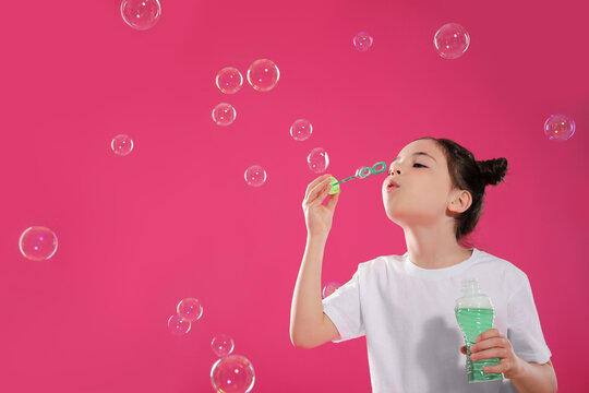 Little girl blowing soap bubbles on pink background, space for text