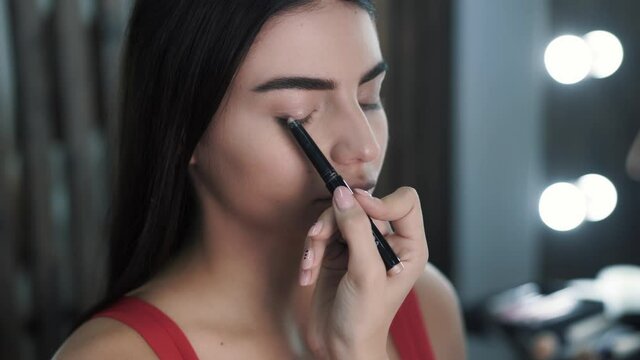 Close-up of makeup artists hand applying brightening shadows to eyelids of caucasian pretty woman model in red dress. Concept preparation for fashion show