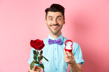 Valentines day. Smiling handsome man asking to marry him, showing engagement ring and red rose,...