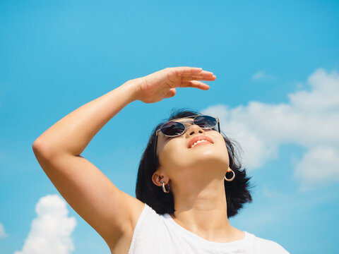 Woman in summertime. Smiling beautiful Asian woman short hair wearing sunglasses and white sleeveless shirt looking up and shading eyes with her hand on blue sky background on sunny day in summer.