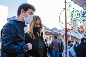 latino hispanic man wearing medical masks with his partner a latina woman at a city fair, looking at handicrafts.