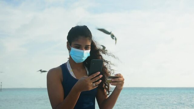 young woman shopping  in mobil phone on  the  beach white covi mask  , seagulls behind in the background