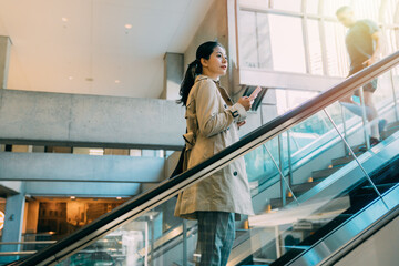Young businesswoman in spring coat holding mobile phone in hand and climbs escalator. Business...