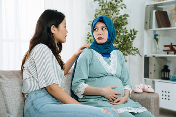 Worried muslim pregnant woman feel stressed and support from asian korean friend in living room at...