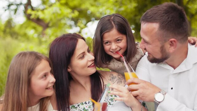 Smiling father, mother and two cute daughters drinking fresh juice at green garden while having summer picnic. Concept of family. Smiling family spending cheerfully free time on nature.