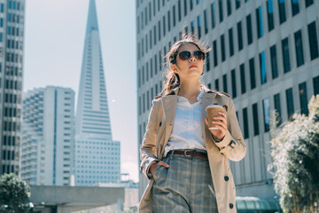 Young beautiful pretty girl walking along street in modern city skyscraper area with hand in pocket...