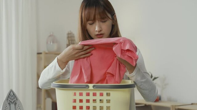 asian girl checking pink shirt over basket found she's messed up burning it with iron. bad house chore experience young lady feels miserable discovering her favorite sweater has unremovable stain