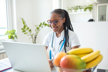 Portrait of young smiling female nutritionist in the consultation room. Nutritionist desk with healthy fruit, juice and measuring tape. Dietitian working on diet plan. - obrazy, fototapety, plakaty