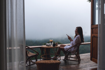 A young asian woman using mobile phone while sitting on balcony with a beautiful nature view on foggy day