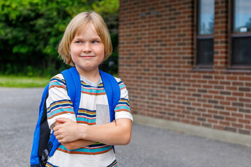 Beautiful happy smiling child near school building at the schoolyard. Little blond student carrying...