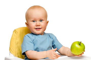The toddler is sitting in a high chair holding a green apple in his hand and smiling, laughing. Healthy nutrition of children with vitamins. Isolate the child of a boy or girl on a white background.