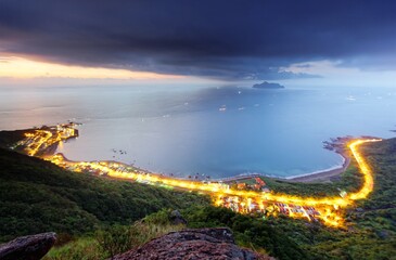 Aerial panorama of a beautiful beach in northern Taiwan under dramatic dawning sky, with a coastal highway along the seashore, lights of fishing boats at sea & an island on horizon in morning twilight