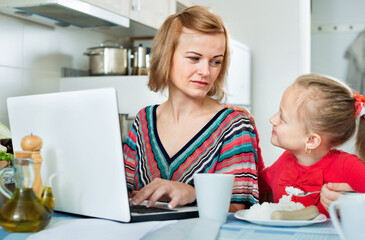 Smiling positive woman sitting with laptop in the kitchen and eating together with her daughter