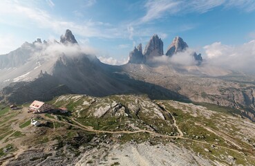 Majestic panorama of vertical peaks of Drei Zinnen (Tre Cime di Lavaredo) & Paternkofel (Monte Paterno) with a mountain hut & hiking tracks wingding on rocky foothills in Dolomiti, South Tyrol, Italy